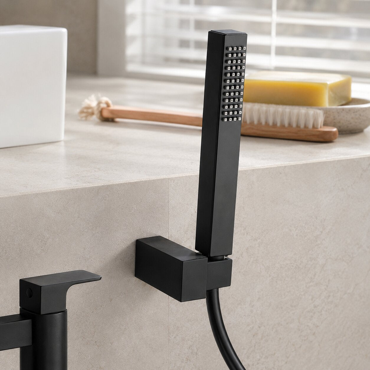 Matte black square handheld shower head with wall-mounted holder and flexible hose, styled beside a modern bathtub in a contemporary bathroom with natural light and premium accessories.
