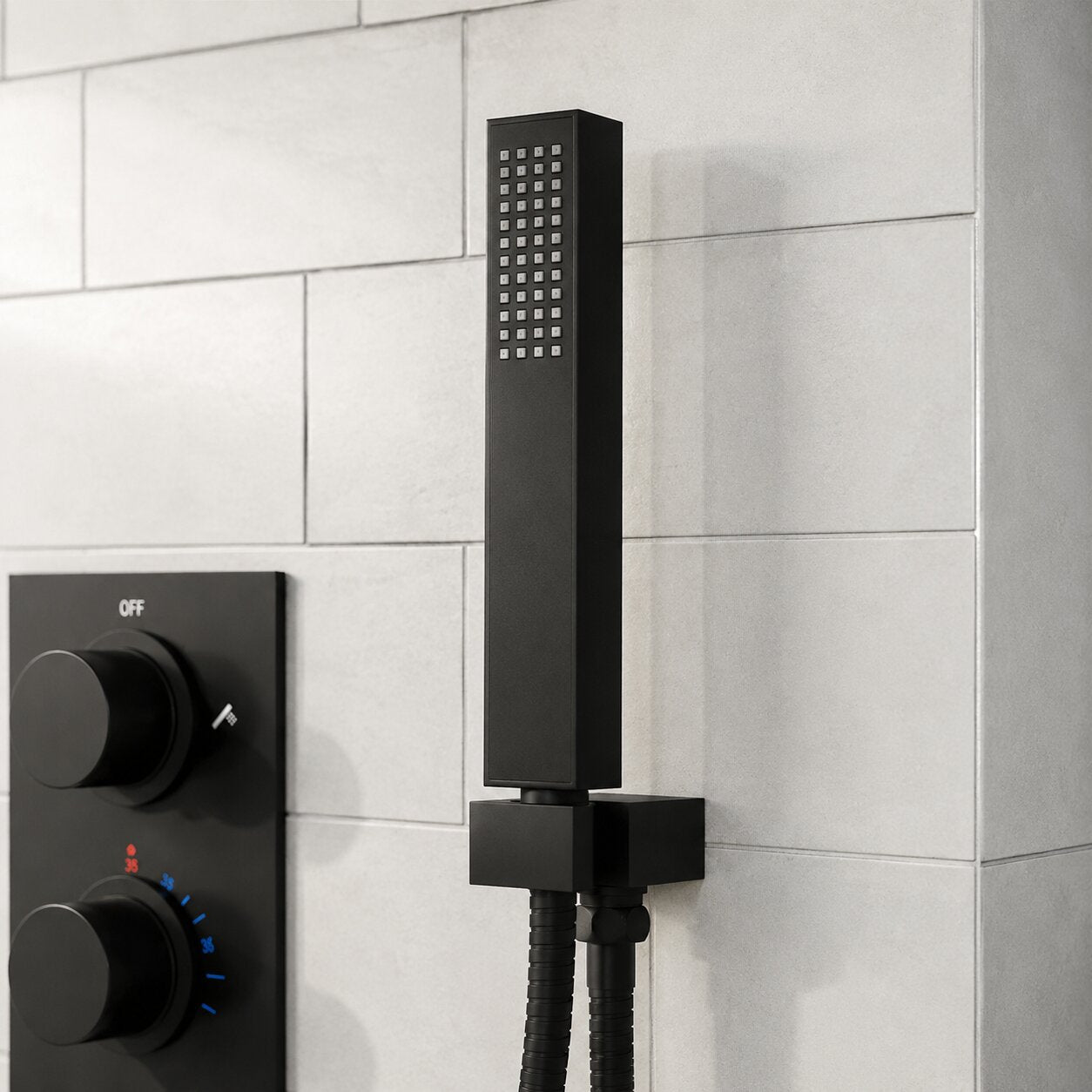 Close-up of a matte black square handheld shower head with matching wall holder and concealed thermostatic shower controls, installed on white subway-effect tiles in a contemporary bathroom.