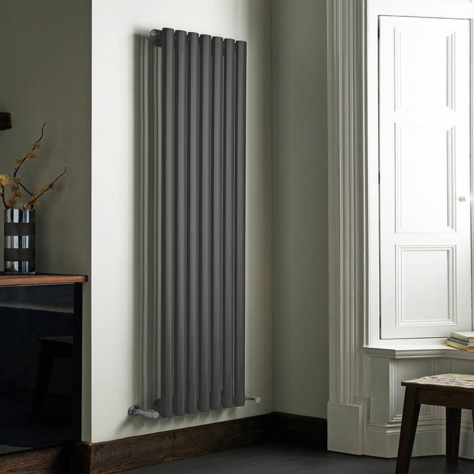 Gray vertical radiator on a white wall in a room with a window and wooden furniture.