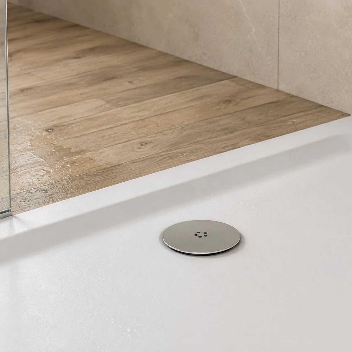 Modern white low profile shower tray featuring a chrome waste cover, frameless glass shower enclosure, wood-effect floor tiles and stone-effect wall tiles in a contemporary UK bathroom.